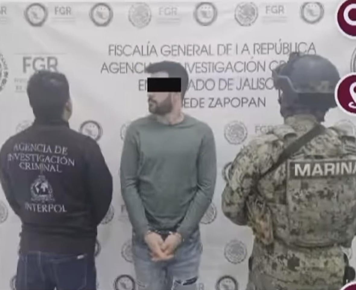 andrew-clark A man in handcuffs is surrounded by someone in an INTERPOL jacket and a soldier in camouflage