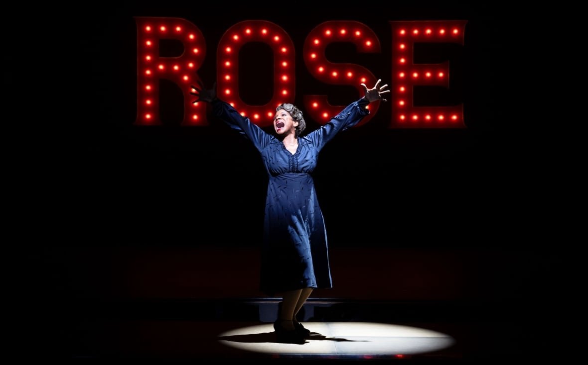 audra-1 A woman stands in a spotlight with her arms spread wide under red lights spelling out the name "ROSE".