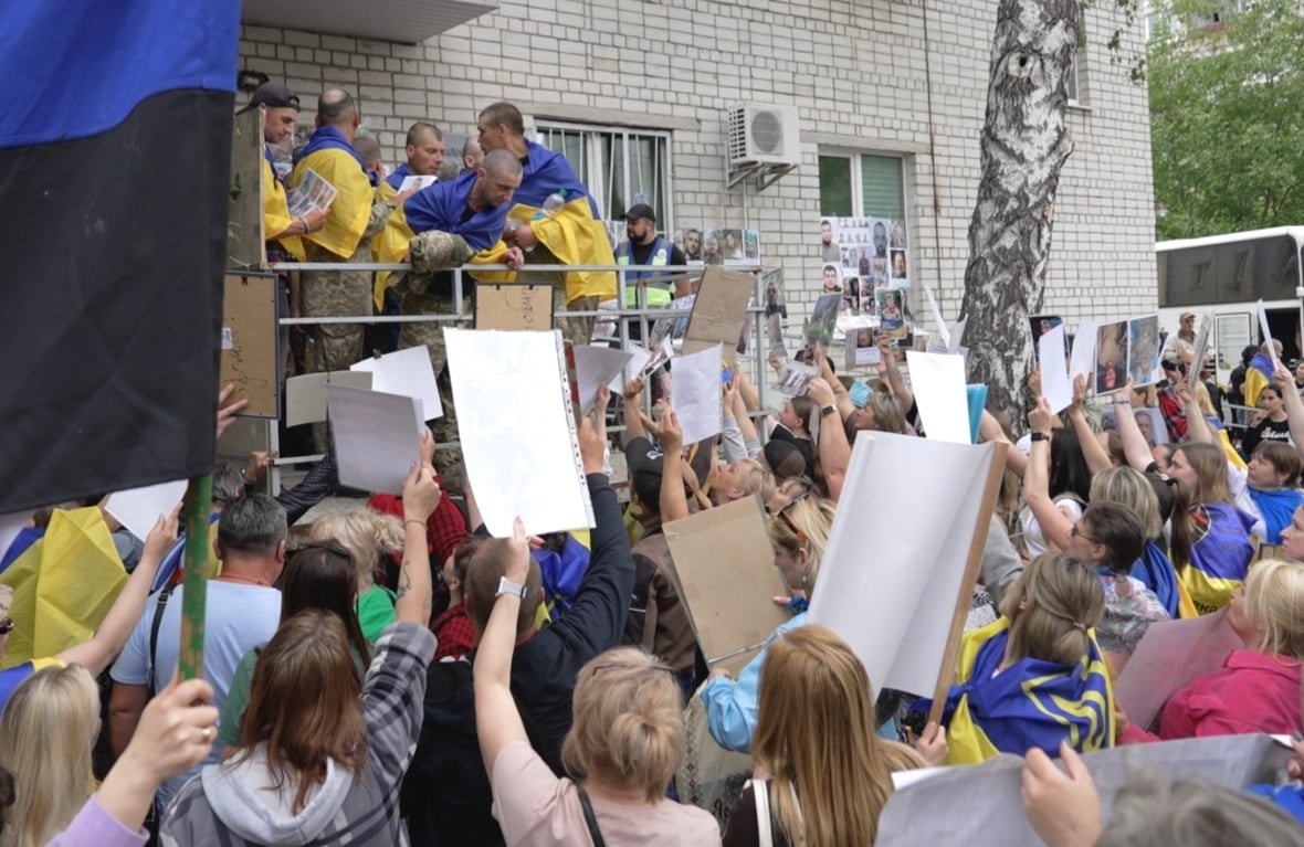 crowd-of-women-holding-photos-of-loved-ones A crowd of people hold up photos and signs as a group of soldiers draped in flags look on.