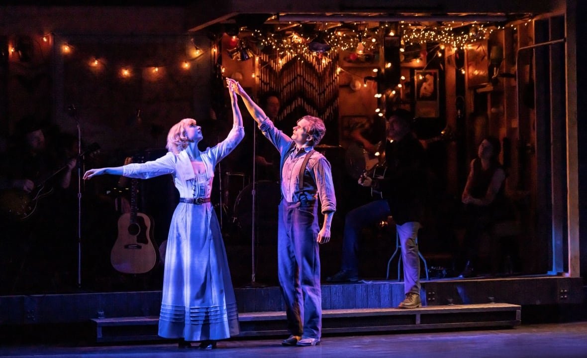 dead-outlaw-1 A man and a woman stand onstage. They each have a hand reached above their heads and towards each other, just touching, and are both looking up towards their hands. They are lit with dramatic blue lighting.