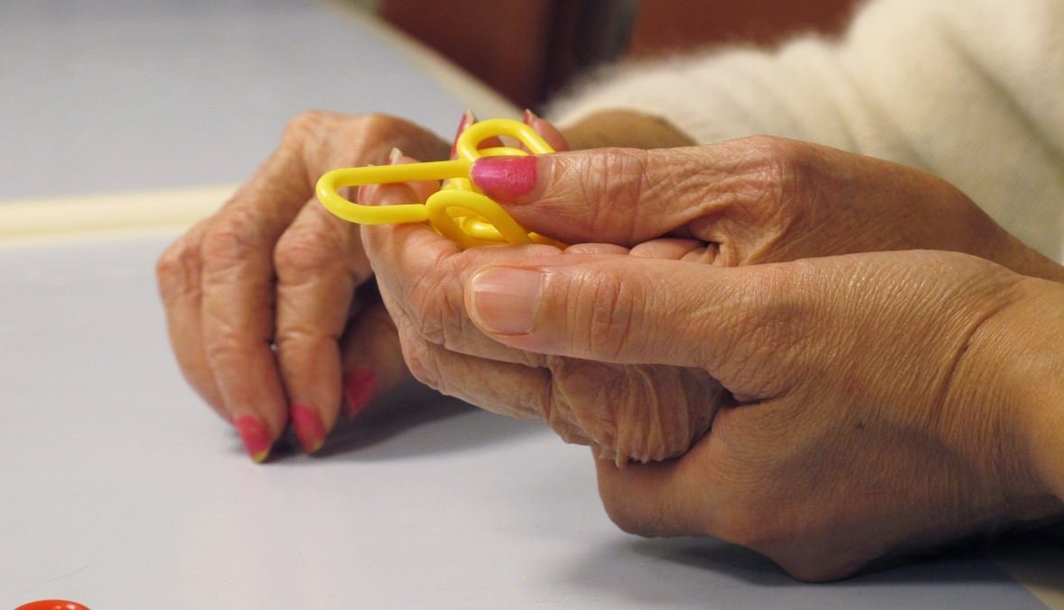 dementia-menopause-symptoms-20240212-1 A close-up of an older person's hands, one resting on a table, the other holding a small yellow toy. Another person is holding one of their hands, cupping underneath to assist them.