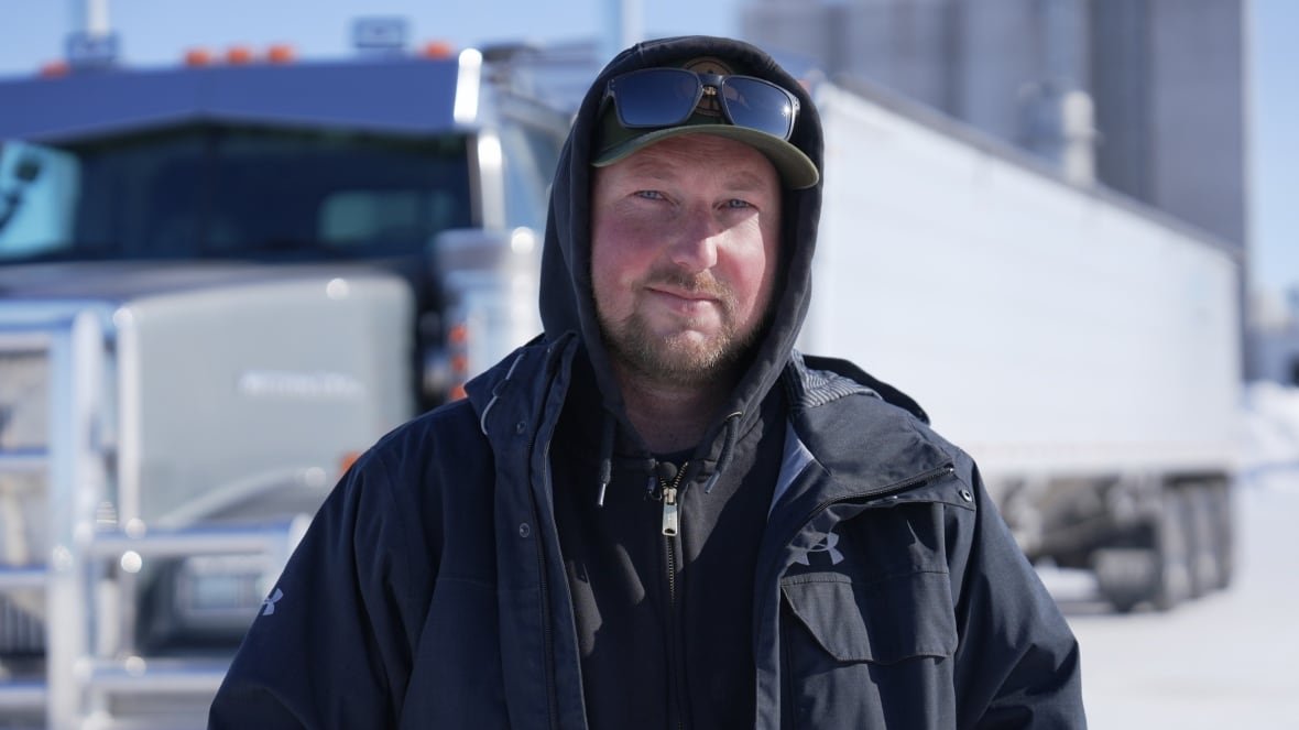 landon-friesen-2 A farmer with a jacket and sunglasses looks into the camera with a semi-truck behind him.