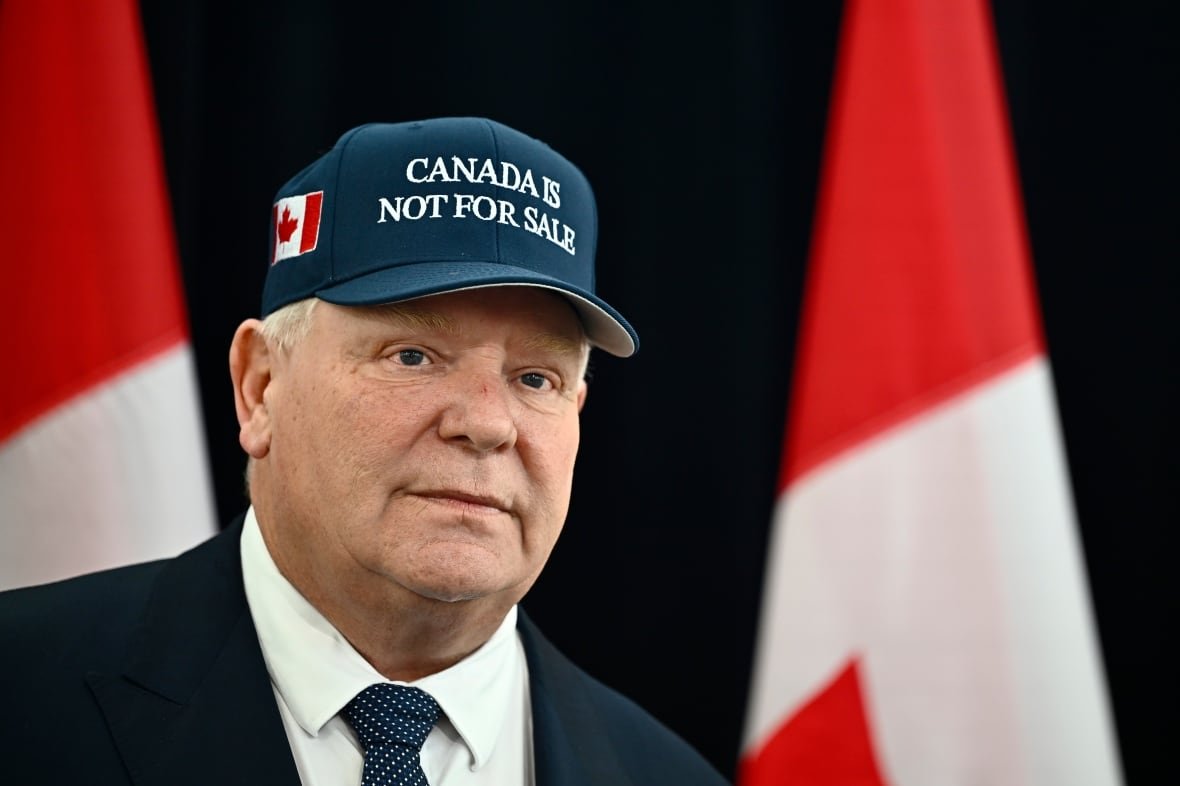 A man wears a navy blue cap, with a small Canadian flag, that reads "Canada Is Not For Sale," as he stands in front of two Canadian flags.