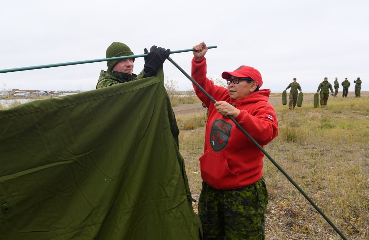 operation-nanook-nunakput-2023 Two people are seen setting up a tent, as a group of military personnel are seen in the background, carrying gear toward them.