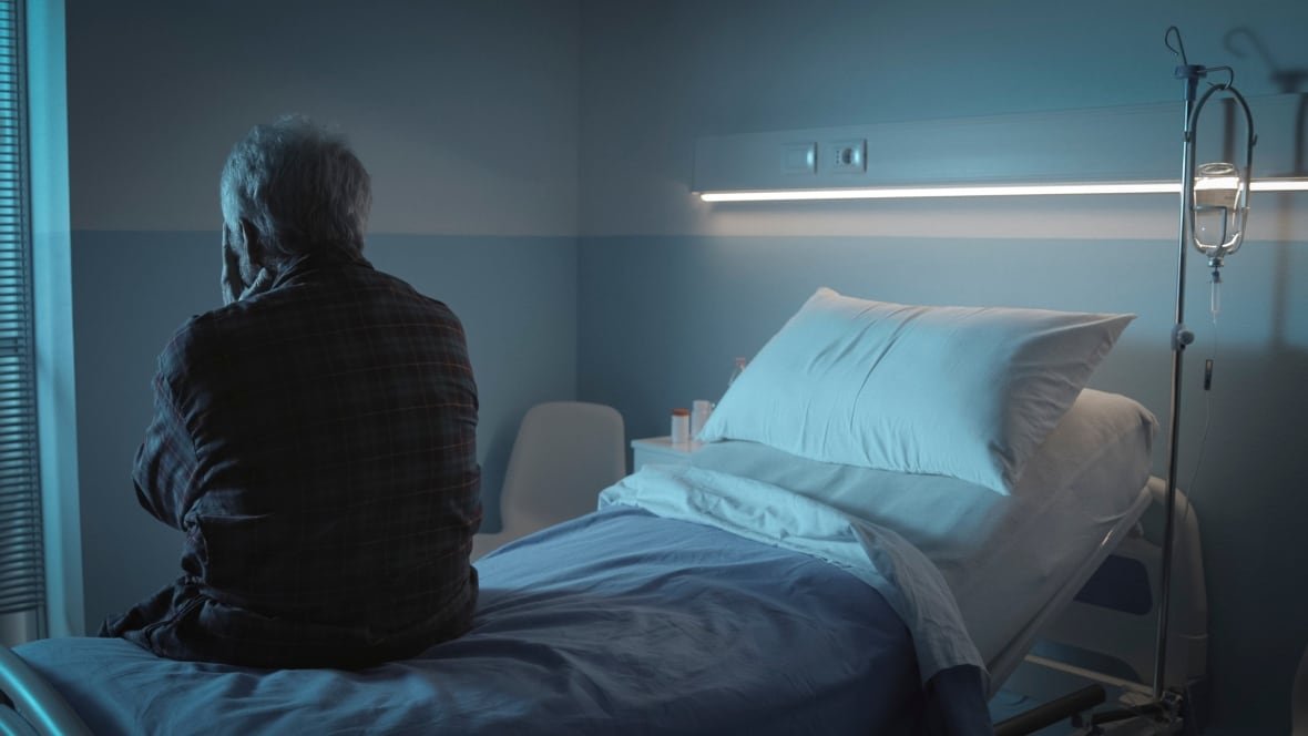 shutterstock-large-file-1 An older person sits on a hospital bed with their back to the camera and their hands over their face. The entire image has a blue cast to it.