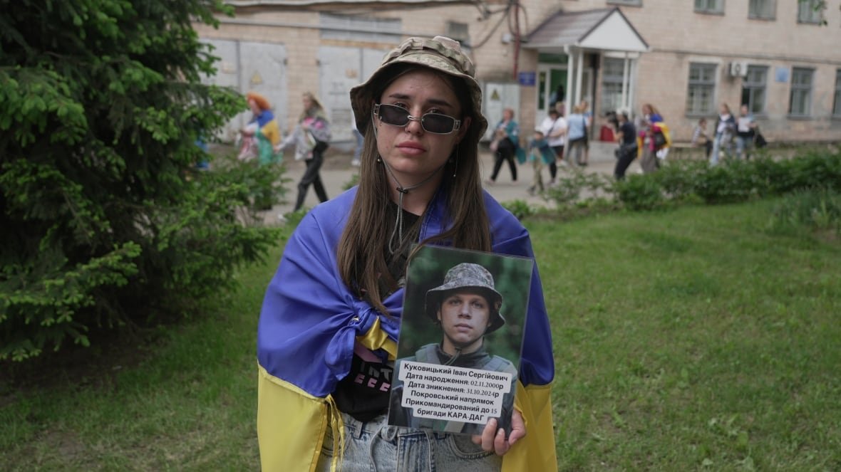 veronika-kulakova A young woman with long brown hair, wearing sunglasses and a hat, holds a photo of a young man.