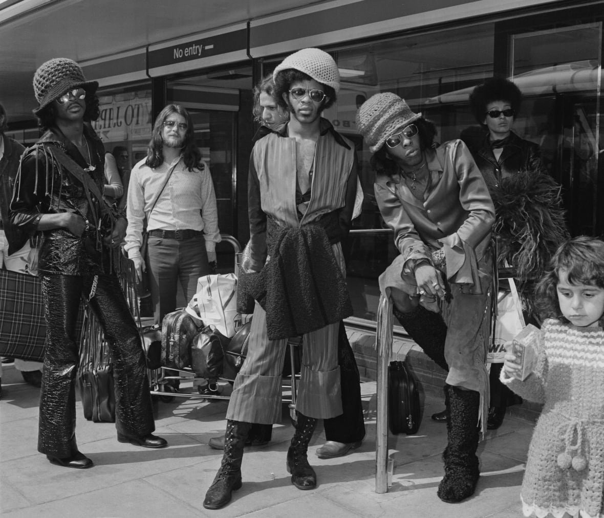 2636208 In this black and white photo, five people pose. The central person is looking at the camera and is slightly in front of the group, with someone else sitting on a table and others standing around. A small child passes by the corner of the image, seemingly unaware of the camera.