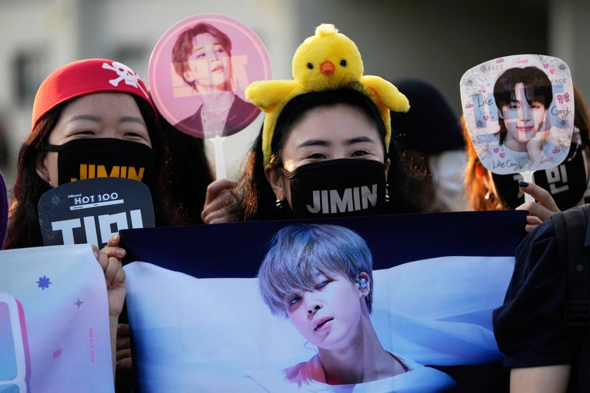 bts-fans-jimin-jung-kook-military-service A crowd of young Korean women, wearing face masks with names written on them, hold up signs and images of young Korean men.