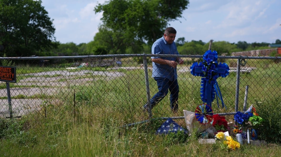 cp174624599-1 A man approaches a memorial near a wire fence, with flowers at the bottom of the fence and a large blue cross attached to the fence itself. The fence crosses a green, overgrown lawn or field, and a structure of some type is visible in the background.