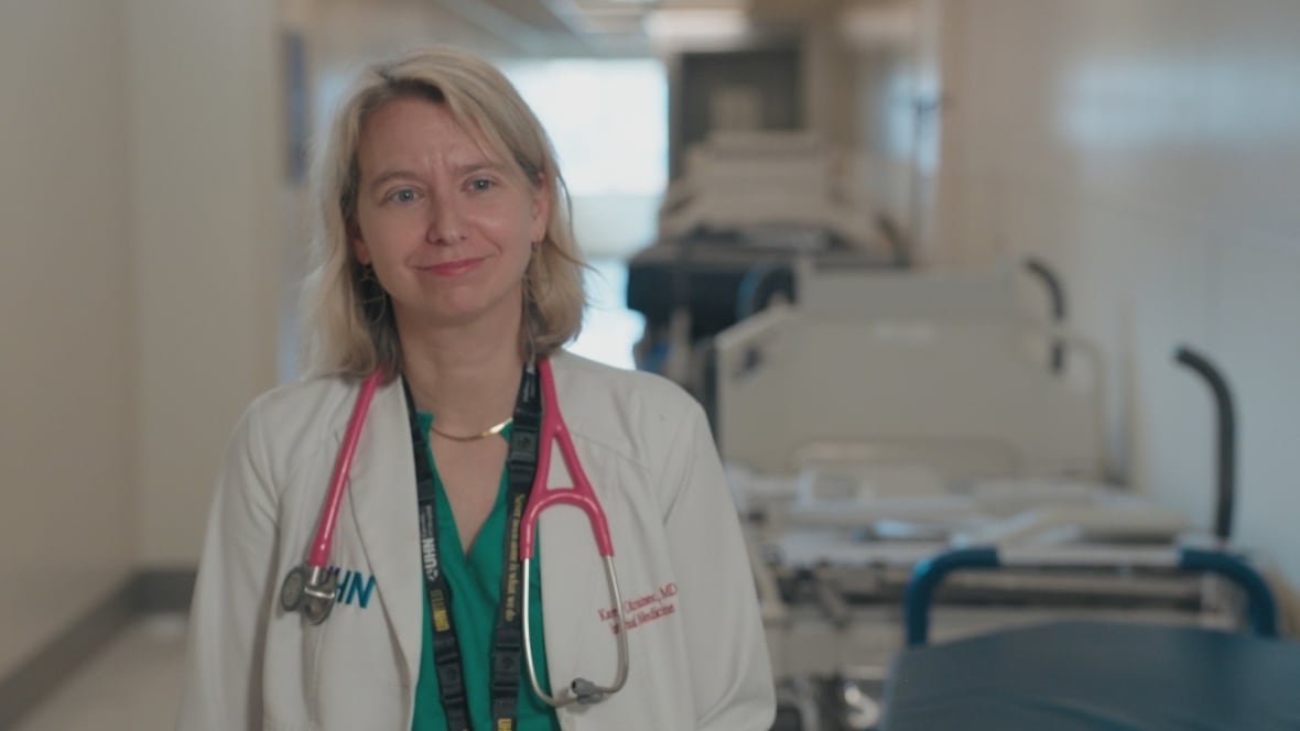 dr-karen-okrainec A doctor with a blonde bob stands in a hospital hallway smiling slightly. There is a pink stethoscope around her neck, over her white coat. The coat has her name - Dr Karen Okrainec, MD - and her hospital network - UHN - embroidered on it.