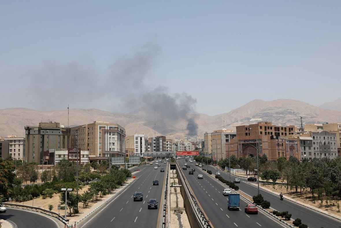 iran-nuclear-opposition-moment-2 Smoke rises from a city in front of a highway, with mountains in the background.