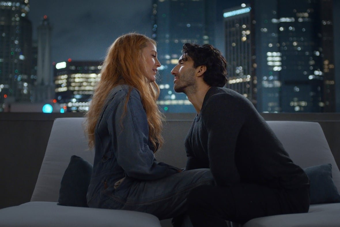 it-ends-with-us A woman with long hair sits on sofa facing a man with wavy dark brown hair, with a city skyline at night seen in the background.