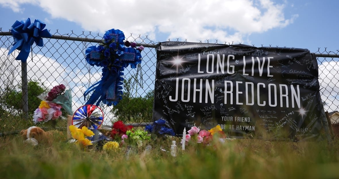 king-of-the-hill-actor-shooting-1 A sign hangs from a fence that says "Long Live John Redcorn" in large letters, with "Your Friend Robert Hayman" underneath. It has been hand-signed many times. Beside it, a large blue cross hangs from the fence, and a number of flowers and flags rest at the bottom of the fence in the grass.