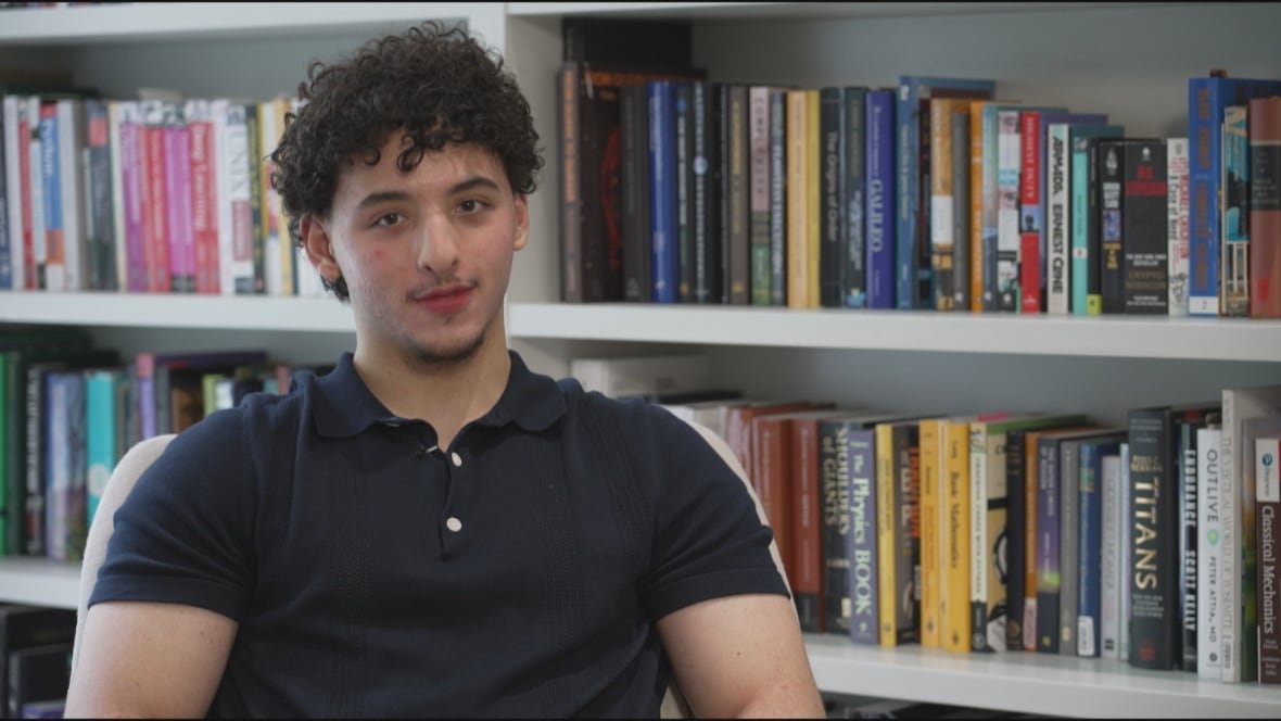 lucas-perri-2 A young man looks at the camera. His hair is curly and he is wearing a blue shirt. There is a bookcase behind him.