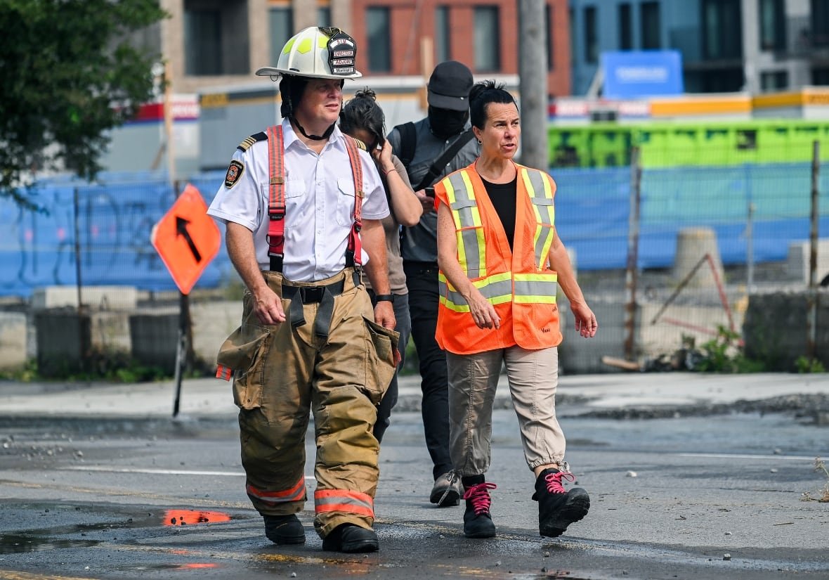 mtl-watermain-break-20240816 A woman wearing a security vest and boots walks next to a fire chief.