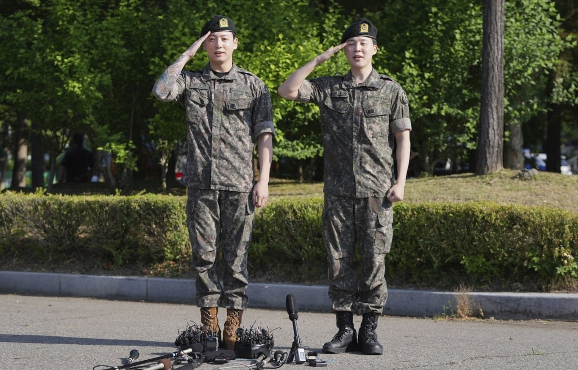 music-suga-bts-returns-1 Two men in army fatigues stand on the pavement outside and salute. There are microphones on the ground at their feet.