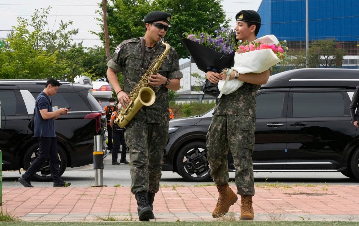 music-suga-bts-returns-2 Two men in army fatigues are pictured. The one on the left is playing the saxophone while the other man looks at him with a smile, holding two bouquets of flowers.