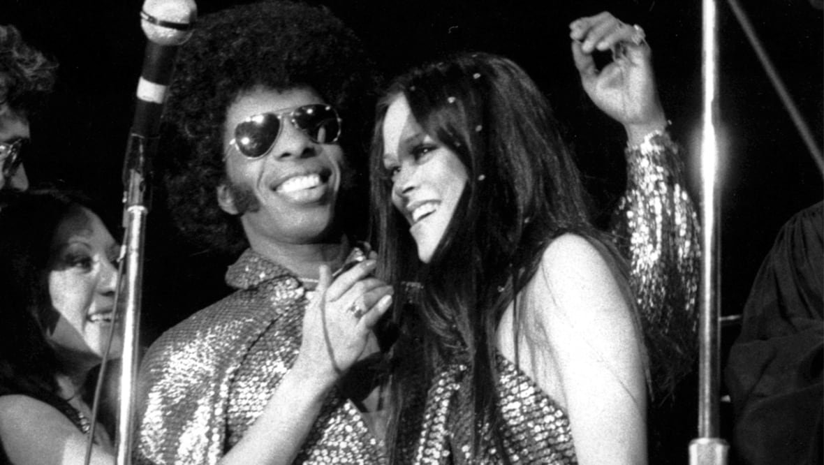 obit-sly-stone A man smiles with his arm around a smiling woman. He is wearing sunglasses and a number of microphones and mic stands surround them. Another woman at the left side of the image reaches towards the first woman's face.