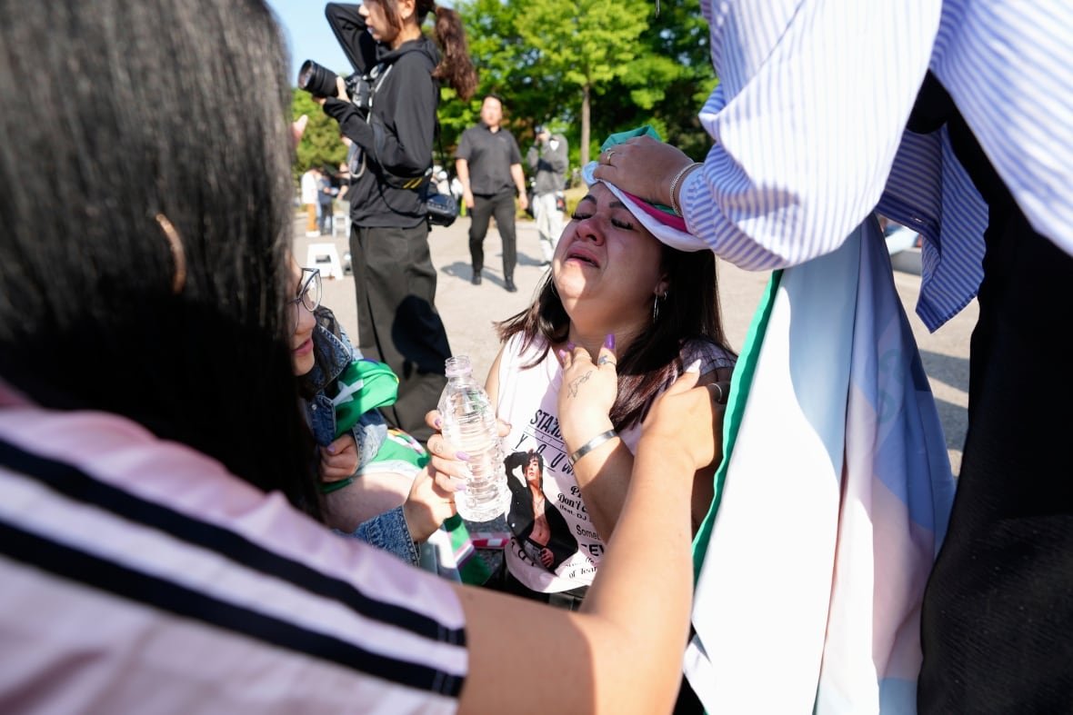 south-korea-bts-fans-jimin-jung-kook A woman in distress sits on the ground, with a bottle of water in her hand, as two other people assist her.