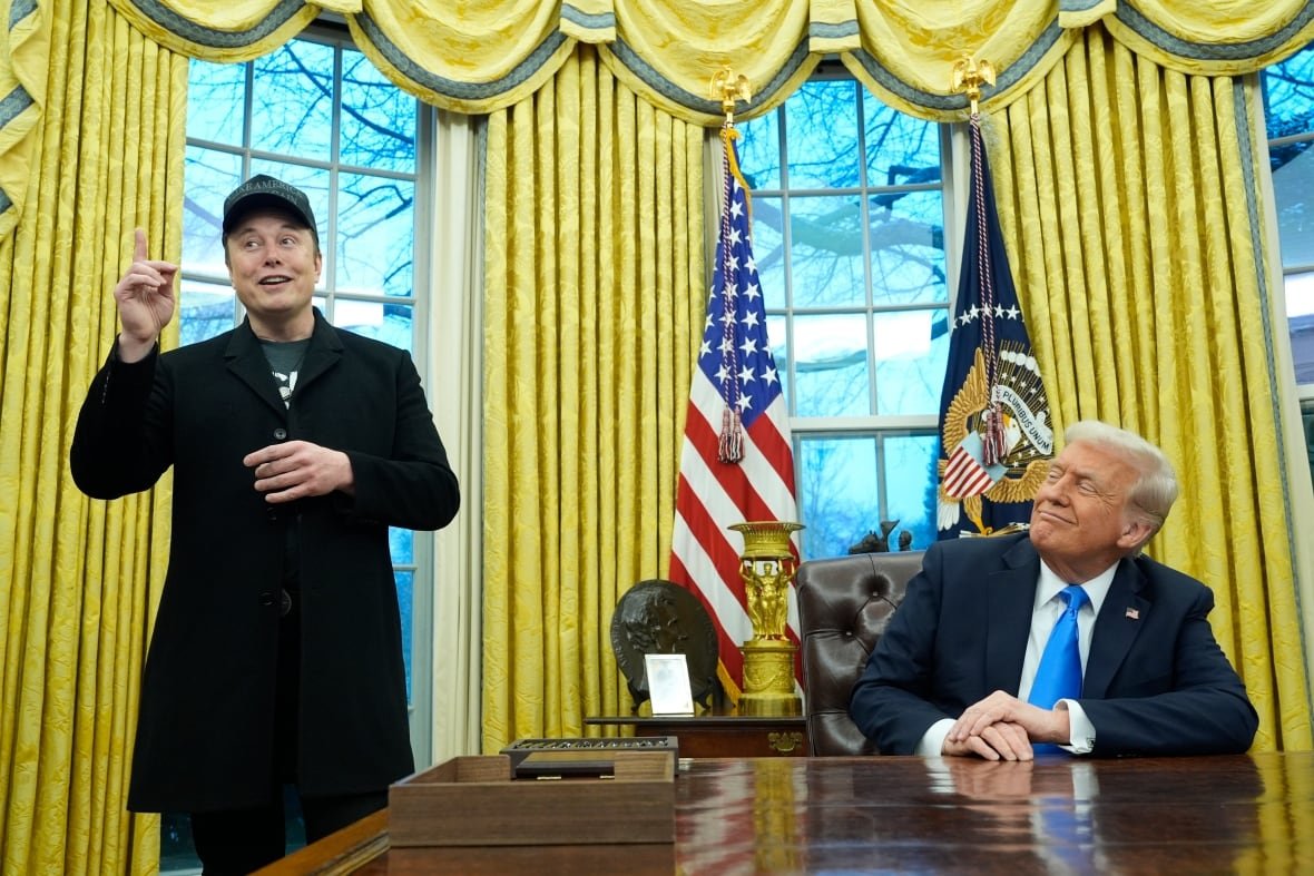 tesla-trump-support-1 A man in a black coat and baseball cap gestures while standing next to a man in a suit seated at a desk. Yellow curtains and two flags are shown in the background.
