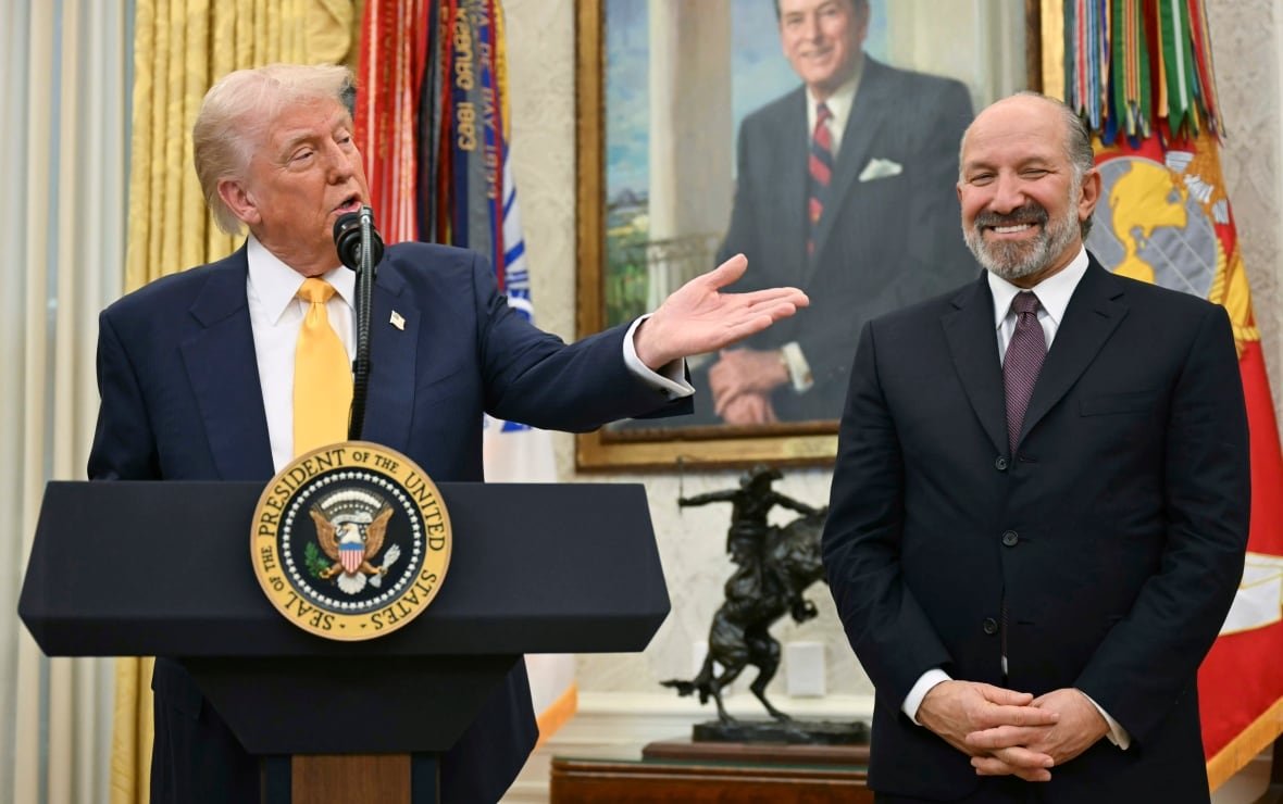 trump-lutnick A blond-haired older man, wearing a navy suit and yellow tie, stands behind a podium and gestures at a bearded grey-haired older man in a dark suit.