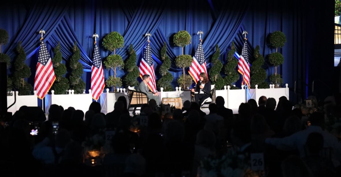 others watching a man and woman sitting onstage in front of several U.S. flags.