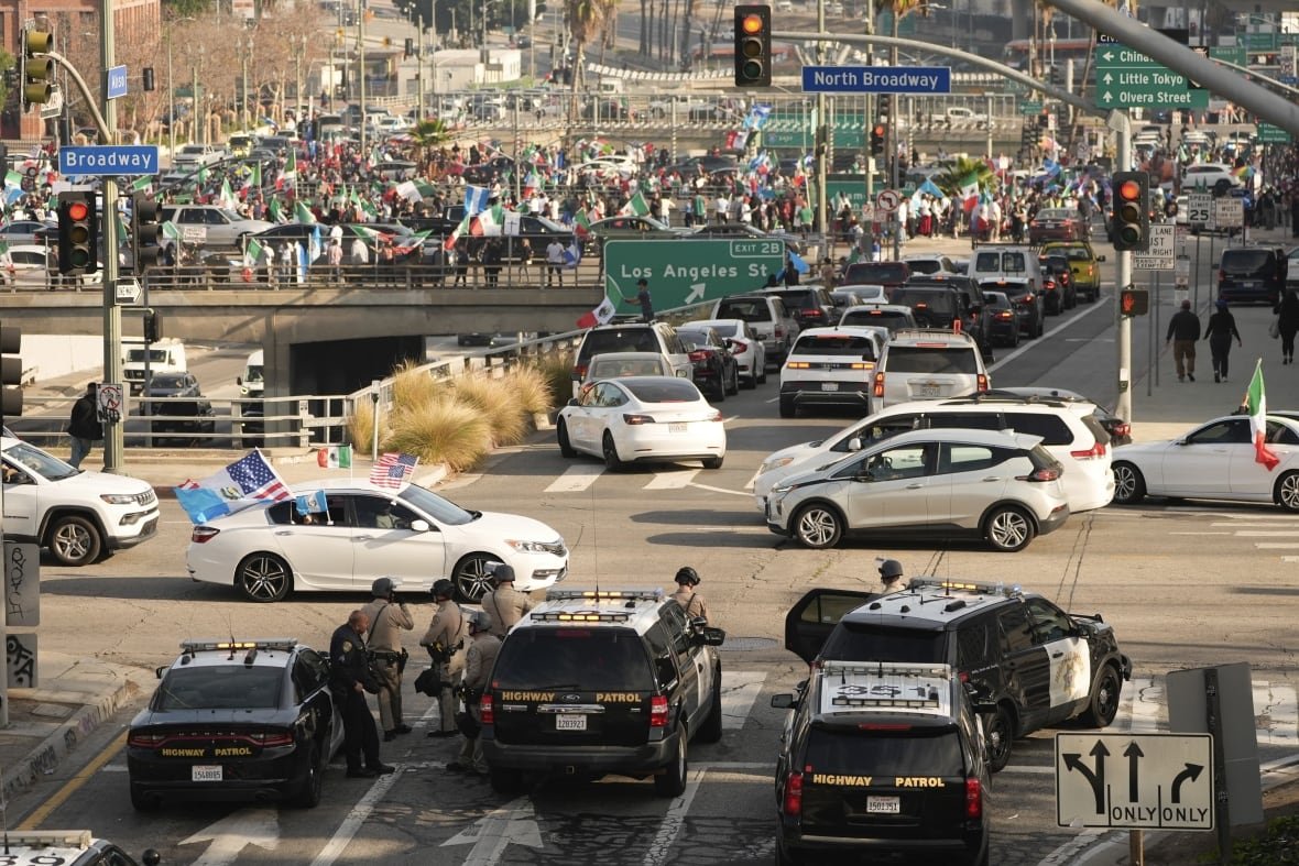 A wide shot from an elevated view shows several police vehicles in the foreground and hundreds of people gathered on a bridge.