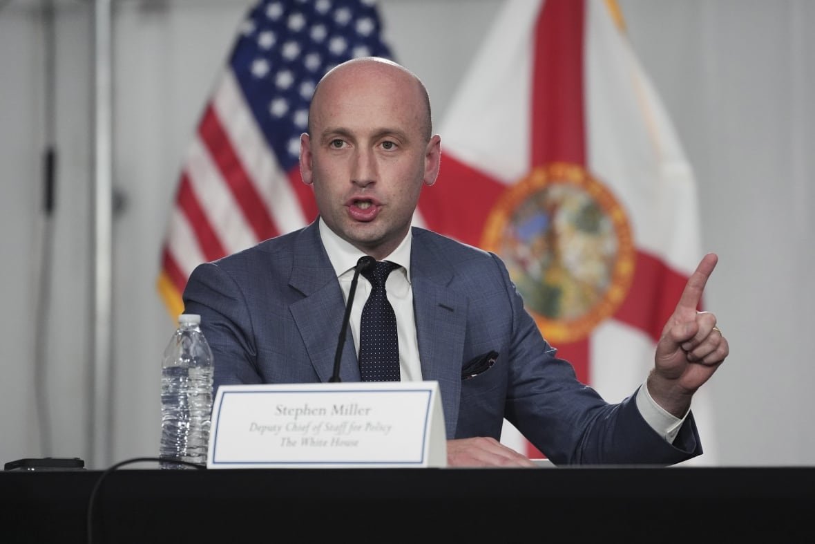 A bald man in a suit and tie holds out his right hand while speaking into a microphone.