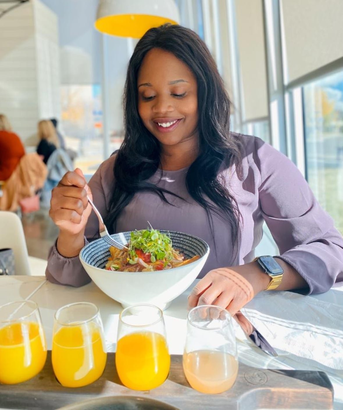 A black-haired woman in a lavendar outfit smiles down at her bowlful of salad in a bright-lit room.