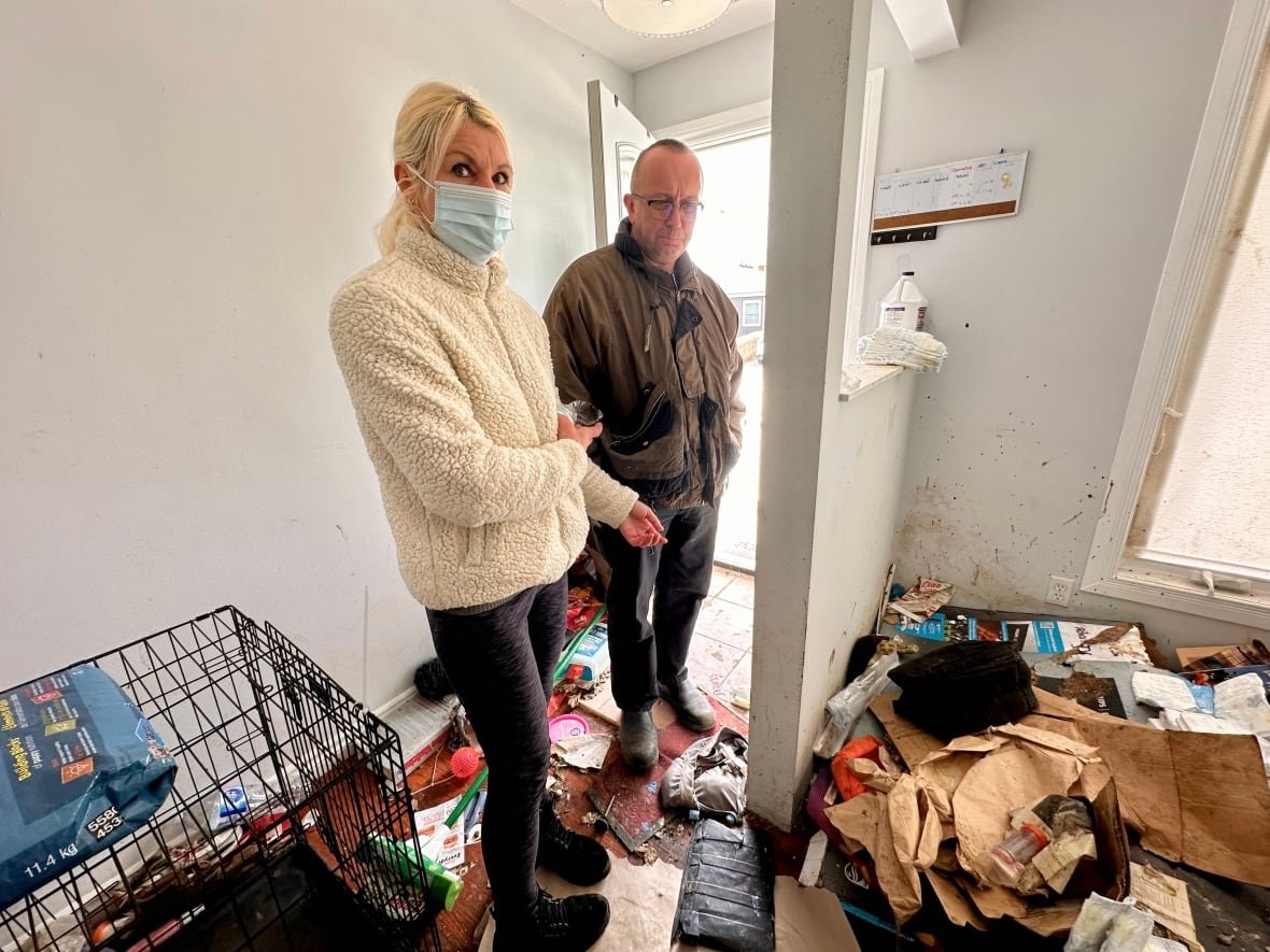 woman in mask stands with man in messy hallway filled with garbage, belongings and a large dog crate.