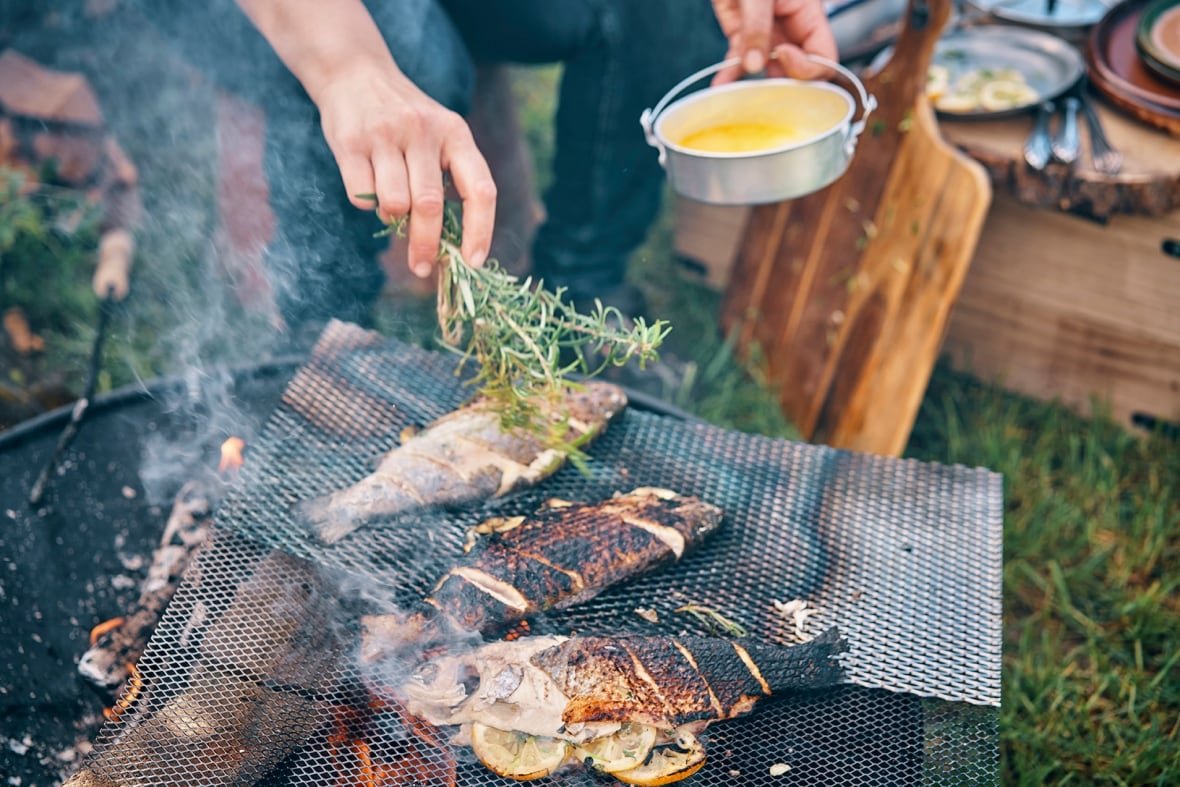 Person preparing fish with rosemary and melted butter over a campfire.