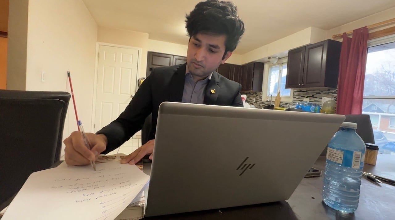 A man writing on a piece of paper with a laptop in front of him.