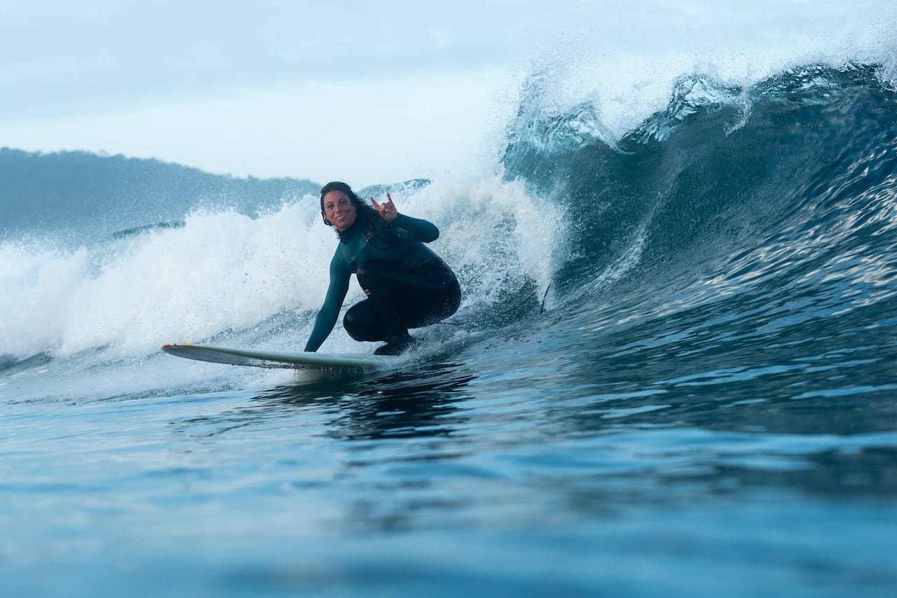 A woman surfing in the winter looks at the camera as she surfs by.