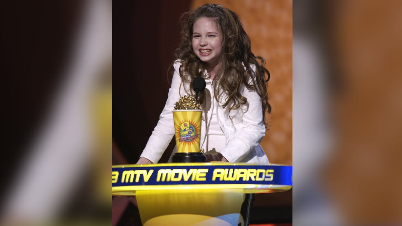 A young girl, with light brown curly hair and wearing a white jacket and white shirt, stands behind a podium with a big smile on her face holding an award statuette resembling a yellow popcorn container with gold popcorn on top.